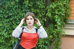 un joven niña en un de moda blanco camisa y rojo parte superior soportes cerca un pared entrelazado con verde hojas y endereza su cabello. foto