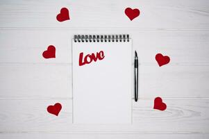 Flatlay composition with blank notebook, pen and red hearts and the word Love on a white wooden background. Love romantic message. Valentine's Day holiday concept. Top view. Copy space. photo