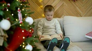 A little boy reads a book while sitting on the sofa near the New Year tree. New Year's mood, a child with a book near the Christmas tree. A boy near a Christmas tree with his book of fairy tales. photo