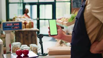 Jeune homme détient téléphone intelligent à boutique vérifier, en présentant isolé écran vert afficher sur mobile appareil. agriculteur et vendeur en utilisant Vide chromakey modèle avec fond technologie. video