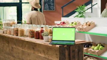 Greenscreen on device in eco store with crates of fresh homegrown products and bulk items in jars on shelves. Laptop showing isolated template with mockup chromakey display. video