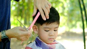 Barber cutting hair of an Asian boy In an open space filled with trees. video