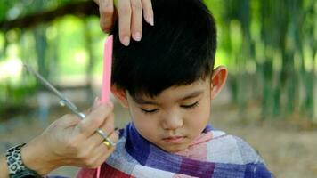 barberare skärande hår av ett asiatisk pojke i ett öppen Plats fylld med träd. video