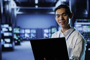 Smiling IT consultant in data center uses laptop to prevent system overload during peak traffic periods. Happy expert in server farm ensuring enough network bandwidth for smooth operations photo