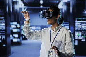 Admin in data center uses virtual reality to prevent system overload during peak traffic periods. Man with VR headset in server farm ensuring enough network bandwidth for smooth operations photo