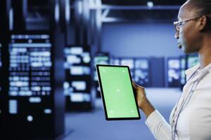 Technician in server room using green screen tablet to future proof network from downtimes and unexpected system failures. Expert with chrome key device ensuring increased data security photo