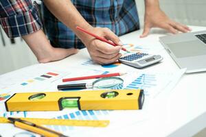 Asian engineer and partner team learn and talk with house plan project blueprint, graph and yellow helmet and engineering construction tools on table work. photo