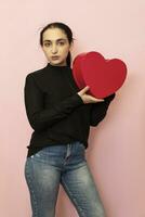 Young middle-eastern woman holds heart shape gift box. Attractive fashion model in casual clothing posing with red present box. Valentines day gift. Love day, anniversary, birthday photo