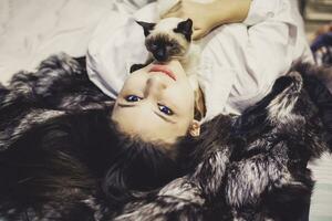 Young middle-eastern woman lying on bed and having fun with her Siamese cat. Pet loving concept. View from above to cute girl playing with cute kitten photo