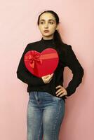 Young middle-eastern woman holds heart shape gift box. Attractive fashion model in casual clothing posing with red present box. Valentines day gift. Love day, anniversary, birthday photo