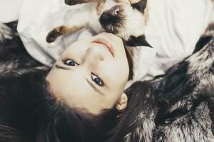 Young middle-eastern woman lying on bed and having fun with her Siamese cat. Pet loving concept. View from above to cute girl playing with cute kitten photo
