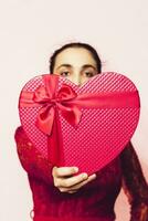 Portrait of lovely woman covering face with giftbox. Valentines day gifts. Beautiful middle-eastern girl receives heart-shaped present box. Love, anniversary, birthday celebration photo