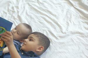 dos pequeño hermanos leyendo un libro. infantil niños acostado en el cama y leer el cuento antes de durmiendo. leyendo un corto historia para niños. medio oriental niños en su cama. foto