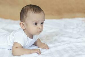 6-8-month-old baby boy lying playfully in bed. Charming 6-7 month little baby in white bodysuit. Baby boy in white bedding. Copy space photo