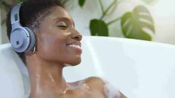 Attractive black woman wearing headphones relaxing with music in foam bathtub. video