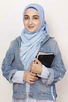 Portrait of cute Muslim businesswoman. Attractive Muslim student holding notepads. Lovely Muslim girl in hijab and denim jacket. Modern, cultural, religious and business concept photo