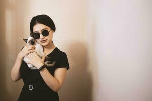 Young attractive woman hugging pussy cat in hands. Cute and glamorous girl in trendy sunglasses posing with her Siamese cat photo