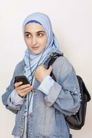 moderno musulmán estudiante niña escucha a música, podcasts, y audiolibros en el teléfono inteligente contento musulmán mujer en hijab escuchando canciones con el auricular foto