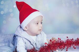 Little funny Santa boy playing with christmas or new year holiday festive garland. 1 year old baby boy in Santa Claus cap. photo