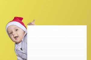 un pequeño chico con Papa Noel gorra participación el blanco papel. Papa Noel bebé chico con un vacío blanco tarjeta. Copiar espacio para tu publicidad y texto foto