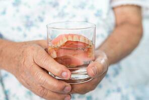 Asian senior woman patient holding and washing denture in water cleanser glass for good chewing. photo