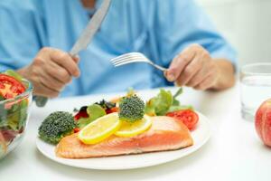 Asian elderly woman patient eating Salmon steak breakfast with vegetable healthy food while sitting and hungry on bed in hospital. photo