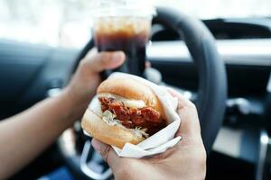 asiático mujer conductor sostener y comer hamburguesa en auto, peligroso y riesgo un accidente. foto