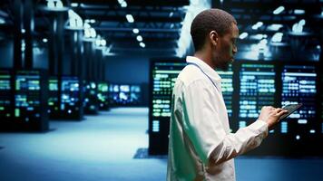 African american IT administrator investigating server clusters delivering web content to online customers. Precise serviceman using tablet to make sure critical systems are working properly photo