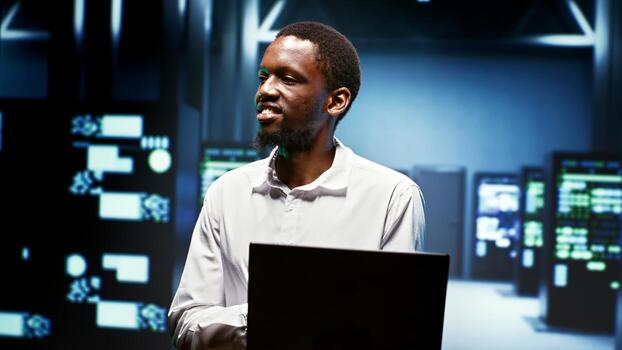 Happy african american employee using laptop to review energy consumption across server cabinets components. Worker making sure data center electronics temperature sensors are operating perfectly photo