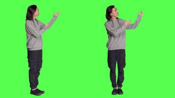 Asian man doing advertisement in studio, pointing at something next to him for new ad slogan. Young person smiling and working on promotional marketing, standing over full body greenscreen backdrop. video