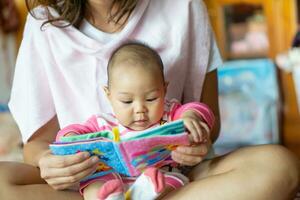 6 meses asiático niña se sentó en el de la madre regazo y leyendo un para niños historia libro. foto