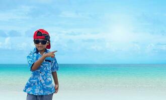 Asian boy wearing sunglasses and a summer outfit is pointing at a free space on the background. photo