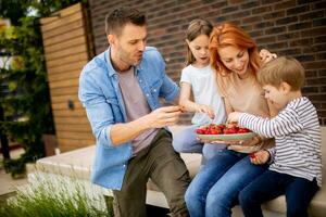 familia con un madre, padre, hijo y hija sentado fuera de en pasos de un frente porche de un ladrillo casa y comiendo fresas foto