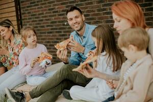 grupo de joven personas y niños comiendo Pizza en el casa patio interior foto