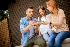 familia con un madre, padre y hija sentado fuera de en pasos de un frente porche de un ladrillo casa y comiendo fresas foto
