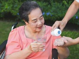 Asian senior woman sitting on wheelchair holding glass of water,  get medical pill from her son. garden, outdoor. photo