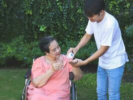 Asian senior woman sitting on wheelchair holding glass of water,  get medical pill from her son. garden, outdoor. photo