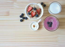 flat lay of  breakfast with oat or granola in white bowl, fresh blueberries, strawberries, a  glass of milk, smoothie blueberries  and yogurt on wooden table. Healthy breakfast concept. photo