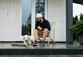 un joven hombre descansa cerca su país casa con un perro. foto