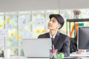 Young Asian entrepreneur sitting at desk with thoughtful facial expression, Businessman thinking about business project at workplace photo