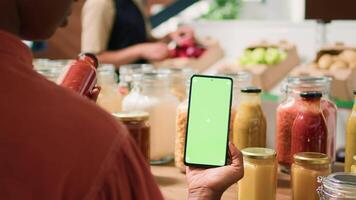 African american client uses greenscreen while she is looking at organic eco bulk products stored in reusable jars. Woman customer checks smartphone with isolated chromakey mockup display. video