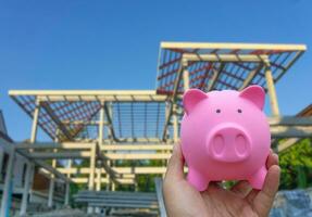 Male hand holding a pink piggy bank on blured construction house background photo