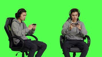 Asian young man plays mobile games over greenscreen, holding smartphone to play online multiplayer competition. Gamer using phone device to have fun with new rpg gameplay on chair. video