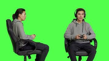 asiatique homme en utilisant manette pour jouabilité, séance sur chaise contre écran vert toile de fond dans studio. Jeune adulte profiter en ligne jeu défi avec amis, joueur en jouant vidéo Jeux. video