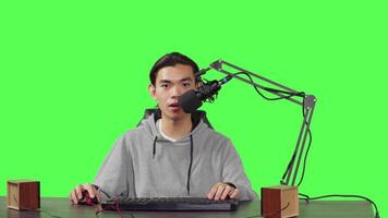 Asian guy does playtime livestream while sitting at workstation and playing online combat video games with his friends. Young man uses microphone to record live broadcast on his desktop pc.