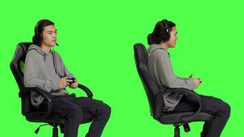 Cheerful person plays video games in studio, having fun with pals on online gaming quest against isolated greenscreen backdrop. Asian man gaming while sitting on office chair, rpg gameplay.