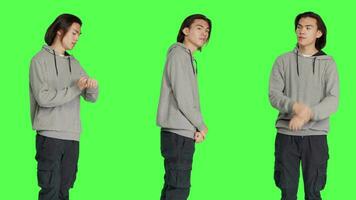 In studio, young adult poses against greenscreen backdrop, looking at wristwatch and appearing agitated. Unhappy and dissatisfied asian person is impatiently awaiting for stuff. video
