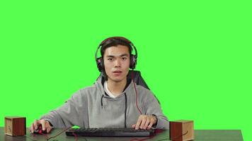 POV of asian guy enjoying computer games at desk while positioned against greenscreen backdrop. Player has fun with friends online in gaming tournament, using headphones at desktop. video