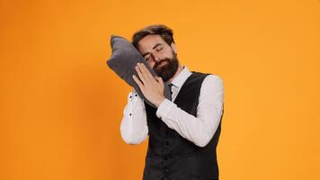 Exhausted waiter taking nap on pillow, sleeping while he is standing over yellow background. Sleepy overworked staff member falling asleep on camera, advertising burnout in catering industry. photo