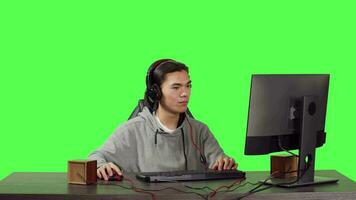 Asian adult being on pc at workstation, playing online rpg video games with his friends. Young man having fun with new web contest, talking to people on headset over greenscreen backdrop.